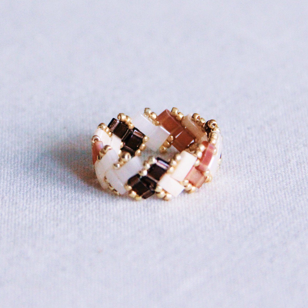 Anillo tejido con cuentas Tila cuadradas en colores marrón, beige y blanco con patrón de espiga y detalles dorados.