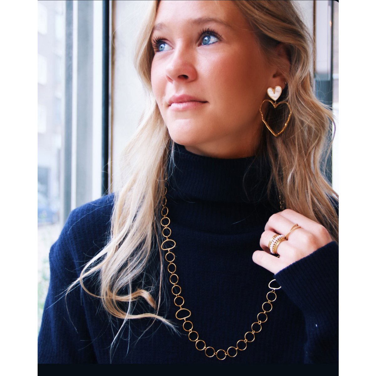 Mujer rubia con jersey azul marino llevando un anillo ancho de perlas combinado con otros anillos dorados.