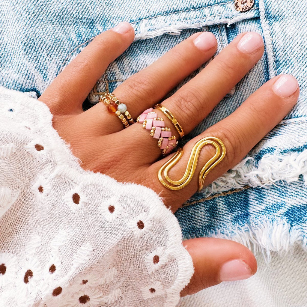 Mano de mujer con anillo XL dorado de forma curva combinado con anillos de cuentas y camisa de encaje.