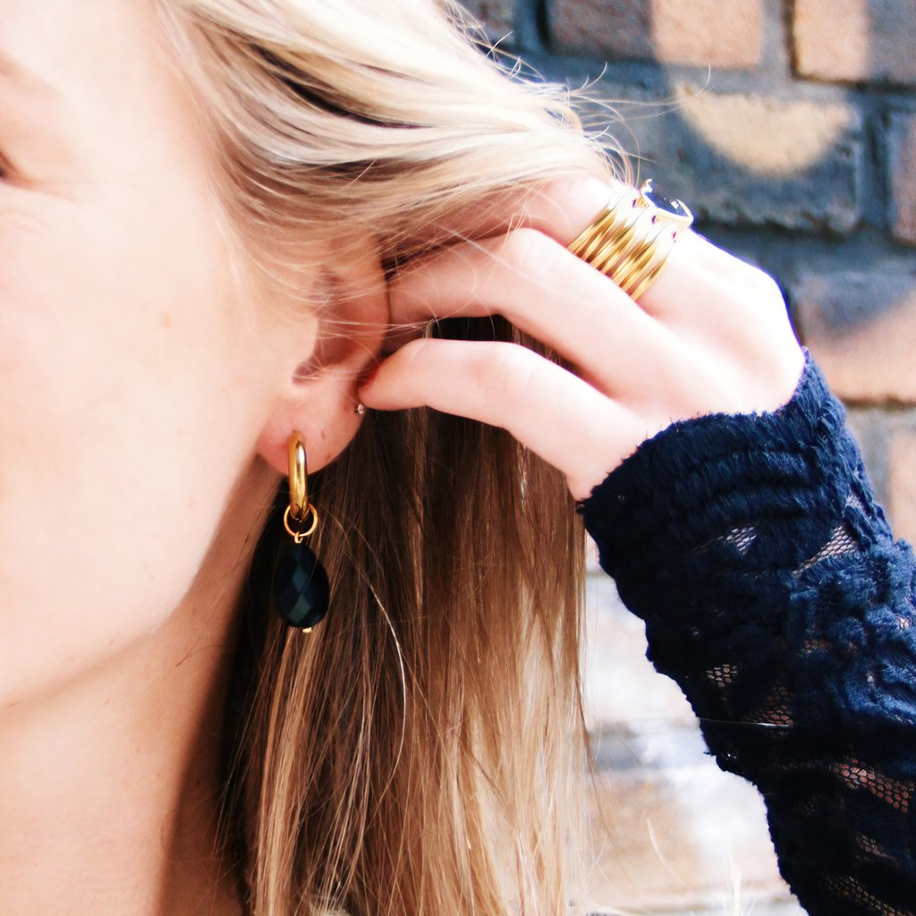 Mujer tocándose el pelo mostrando pendientes de gota negra y combinación de anillos dorados.