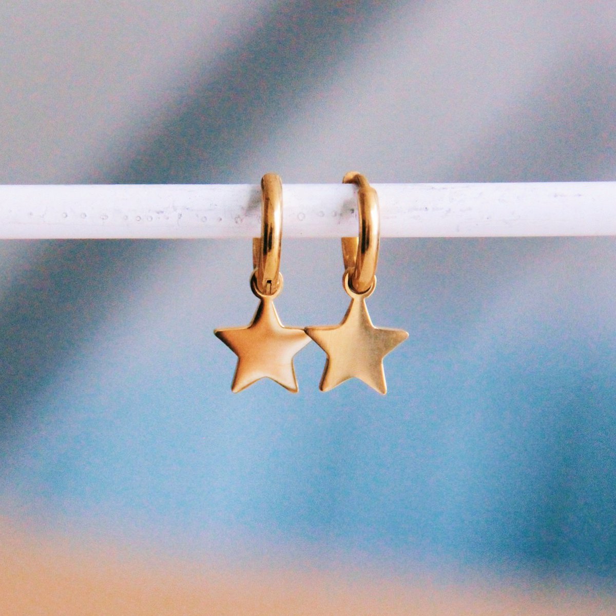 Par de aros pequeños dorados de acero inoxidable con charm de estrella colgante sobre fondo neutro.