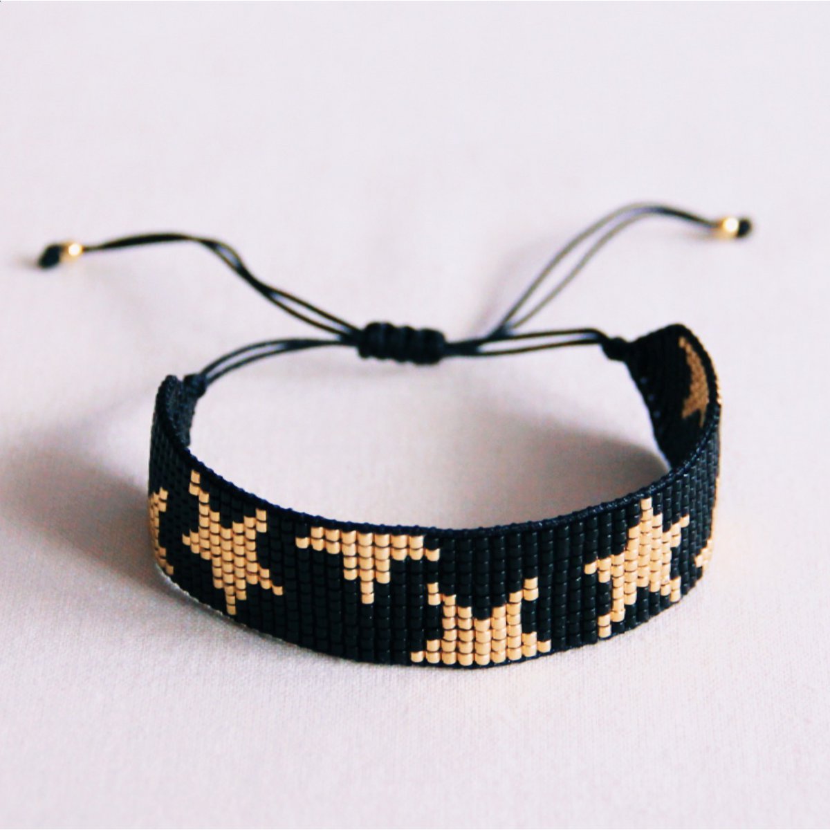 Pulsera tejida de estrellas en negro y oro con cierre de nudos corredizos ajustable sobre fondo blanco.