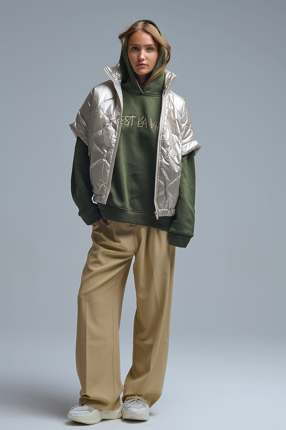 Mujer con look completo vistiendo un chaleco acolchado metalizado de manga caída, sudadera verde, pantalones beige y zapatillas chunky.