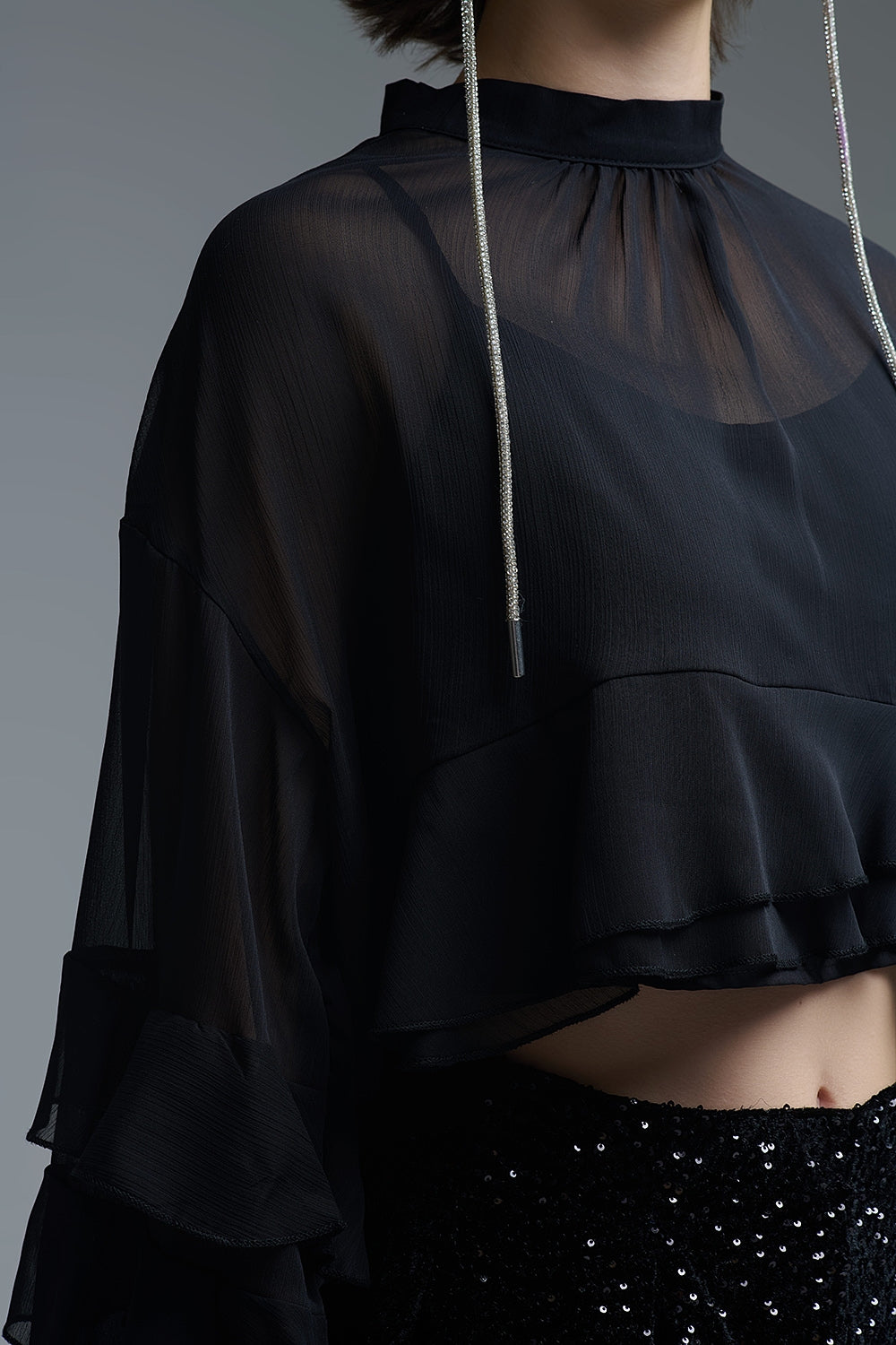 Detalle lateral de los volantes y la textura de gasa de una blusa negra estilo capa corta.