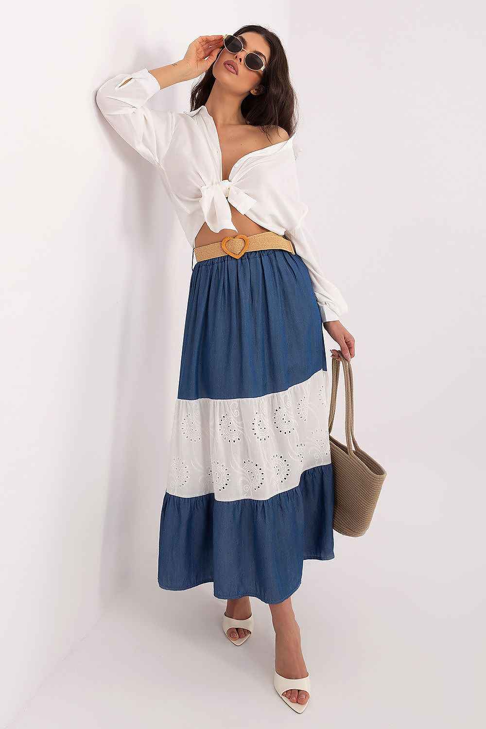 Mujer posando con falda larga de paneles en azul denim y blanco con bordado suizo, camisa blanca anudada y cinturón de rafia.