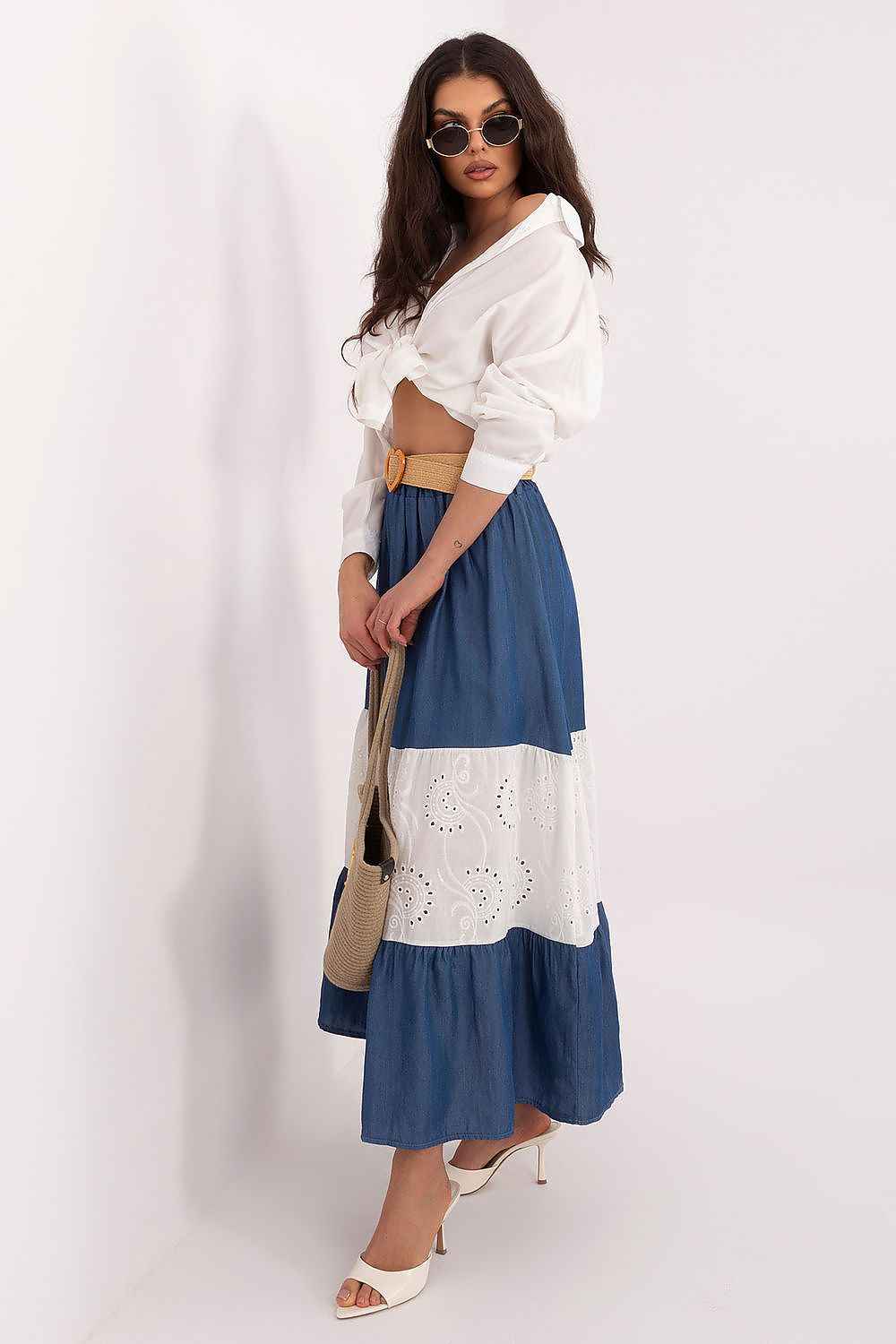 Mujer posando con falda larga de paneles en azul denim y blanco con bordado suizo, camisa blanca anudada y cinturón de rafia.