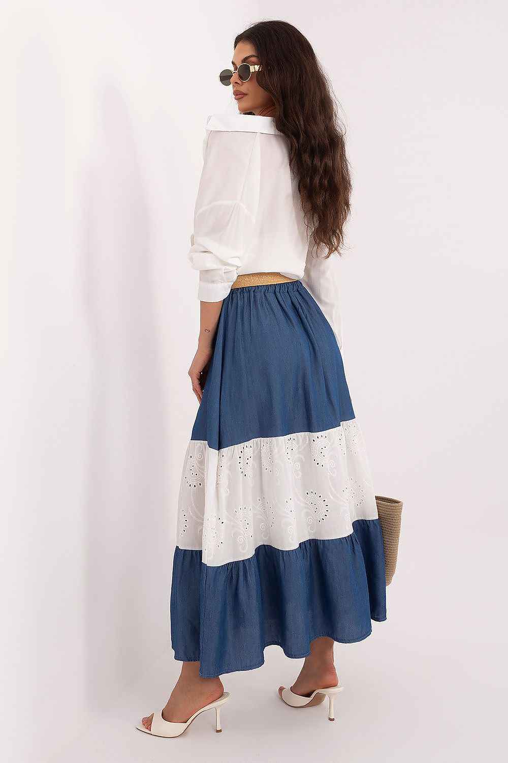 Mujer posando con falda larga de paneles en azul denim y blanco con bordado suizo, camisa blanca anudada y cinturón de rafia.