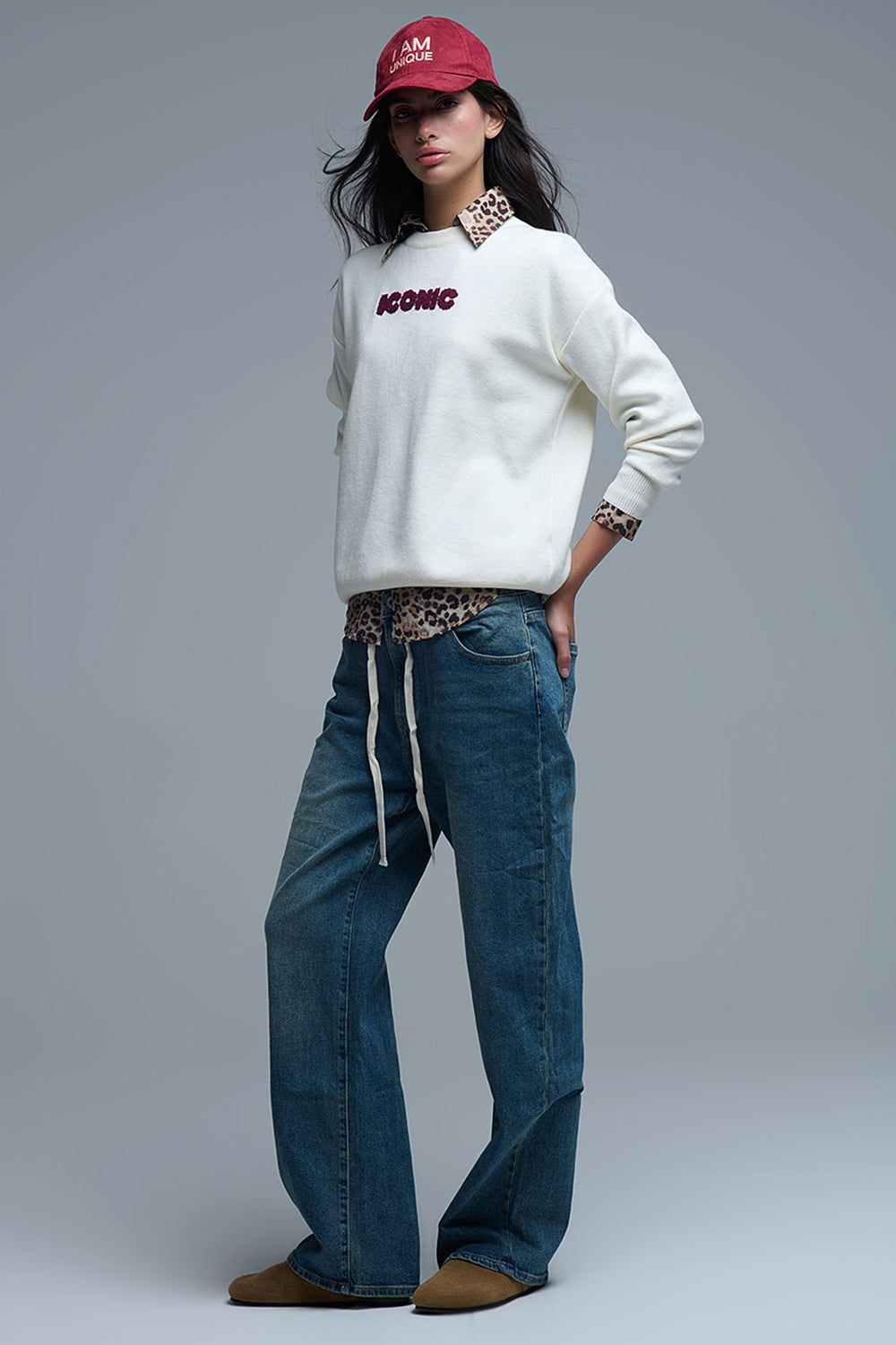 Mujer con gorra y jersey de punto crema "ICONIC" sobre una camisa de leopardo y con vaqueros baggy.