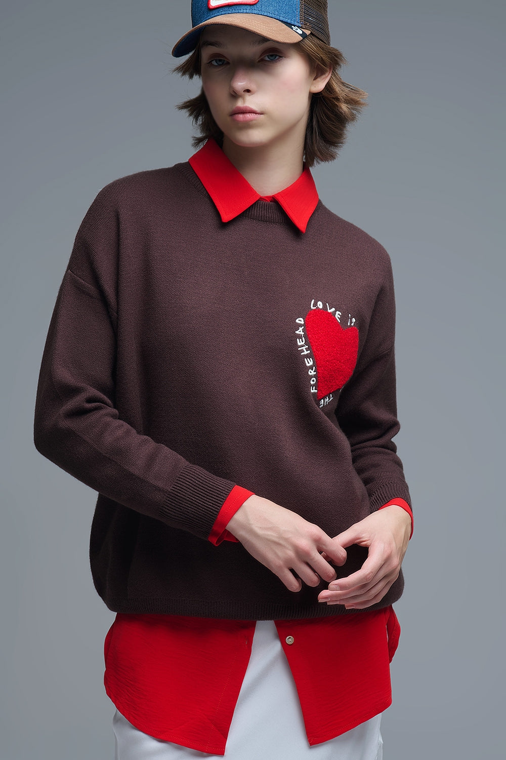 Modelo con look preppy de jersey oversize marrón con un corazón rojo bordado en el pecho, sobre una camisa roja.