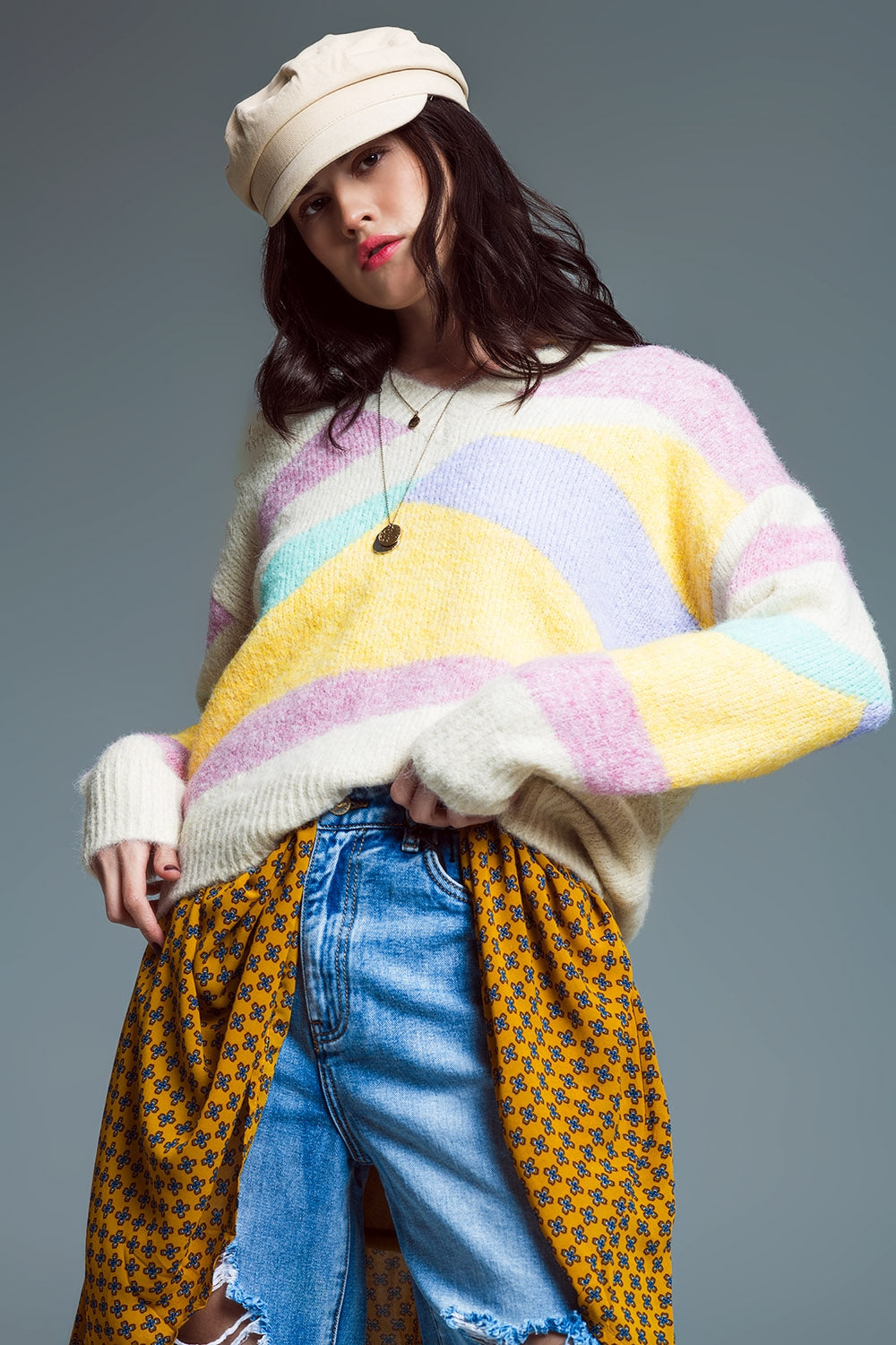 Mujer con gorra beige vistiendo jersey de punto grueso con rayas abstractas en colores pastel y vaqueros rotos.