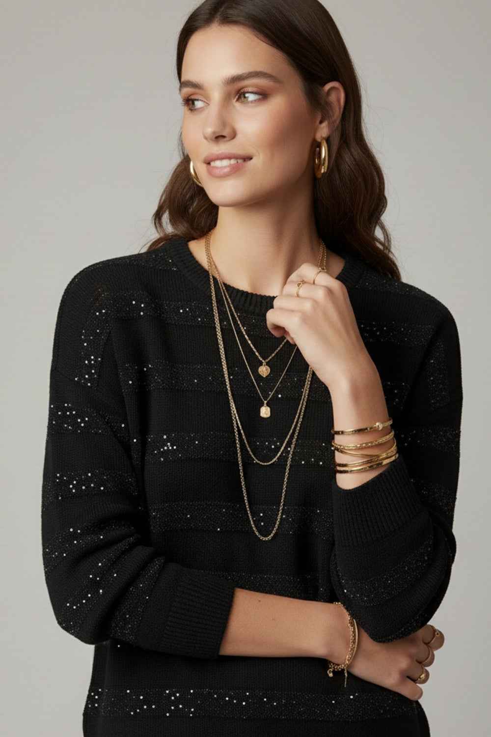 Mujer luciendo jersey de punto negro con rayas horizontales de lentejuelas brillantes, cuello redondo y accesorios dorados.