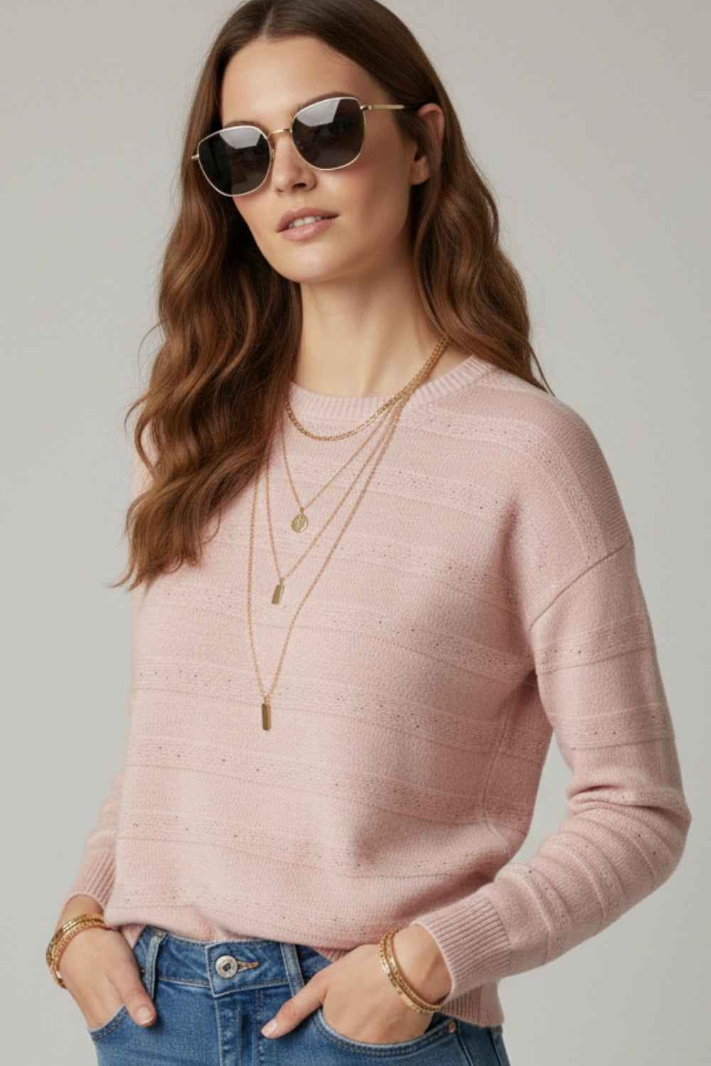 Mujer con jersey de punto rosa empolvado, rayas de lentejuelas, gafas de sol y collares dorados.