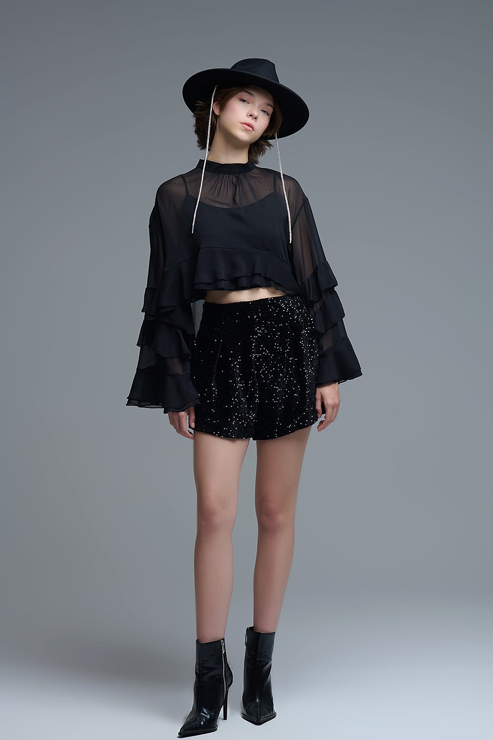 Modelo posando con look de fiesta negro completo: blusa de volantes transparente, shorts brillantes y sombrero.