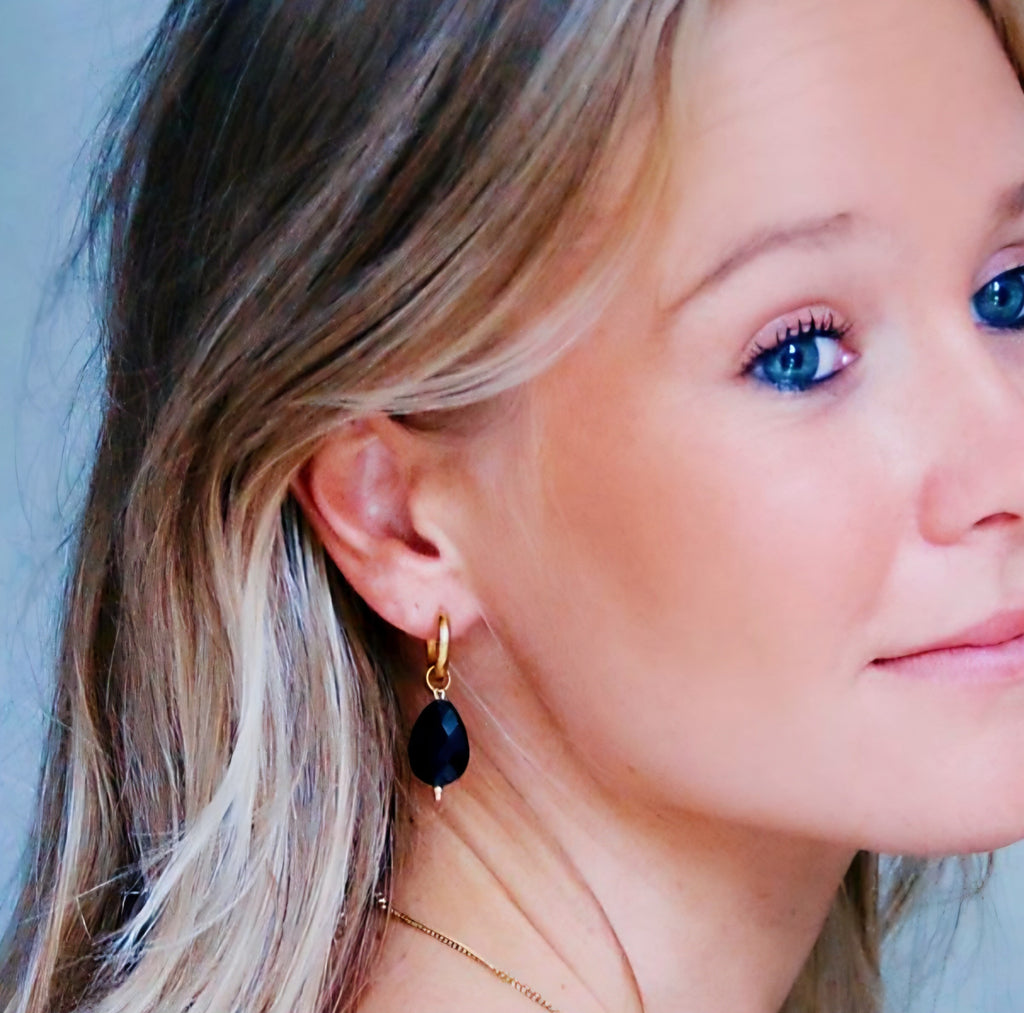 Primer plano de mujer con pendientes de aro grueso dorado y colgante de gota negra facetada.