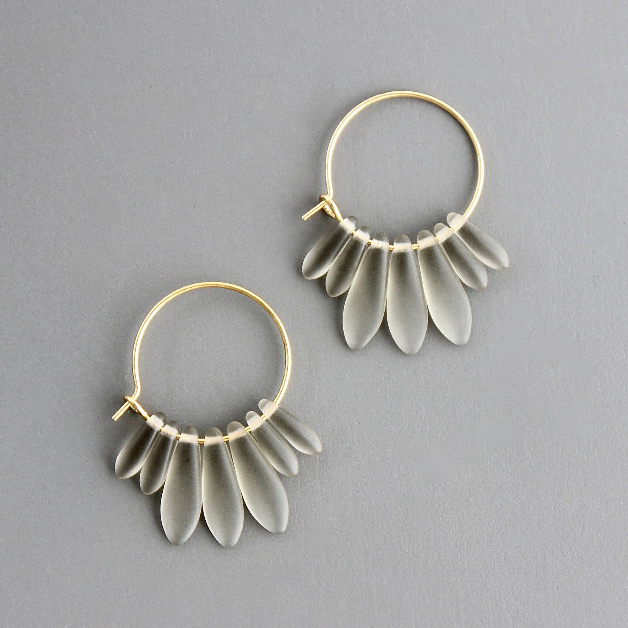 Pendientes de aro dorado con cristales checos mate transparentes, diseño artesanal de David Aubrey sobre fondo gris.