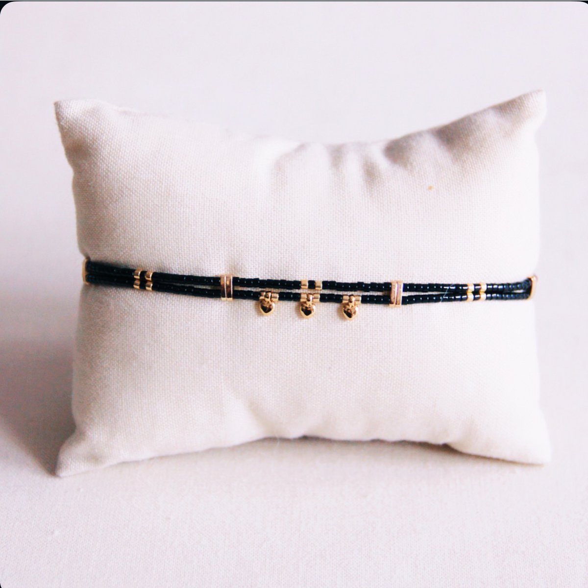 Pulsera de doble hilera de cuentas Miyuki negras con tres mini charms de corazones dorados colgantes sobre cojín.