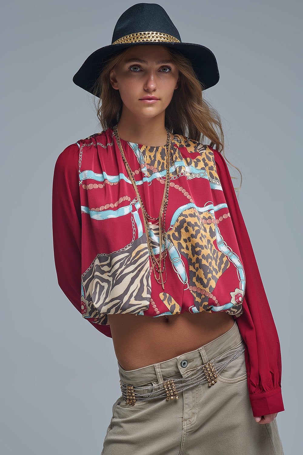 Mujer con sombrero y top corto rojo con estampado de pañuelo, cadenas y leopardo, y pantalones beige.