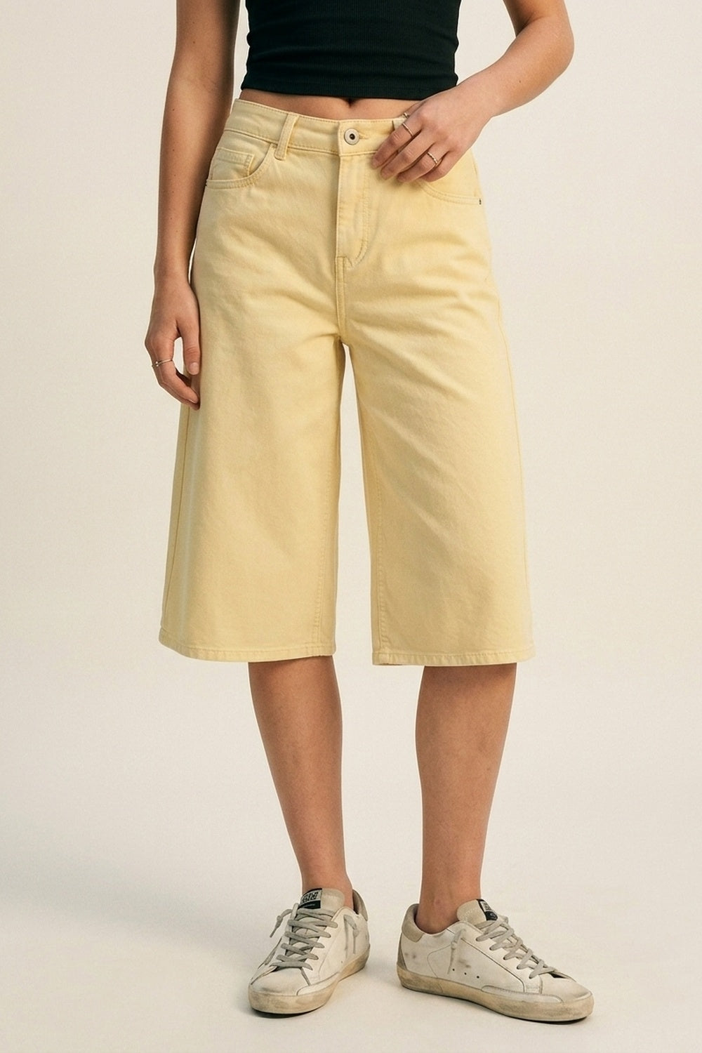 Bermudas vaqueras tipo culotte en color amarillo pastel de tiro alto, combinadas con top cropped negro y zapatillas deportivas envejecidas.