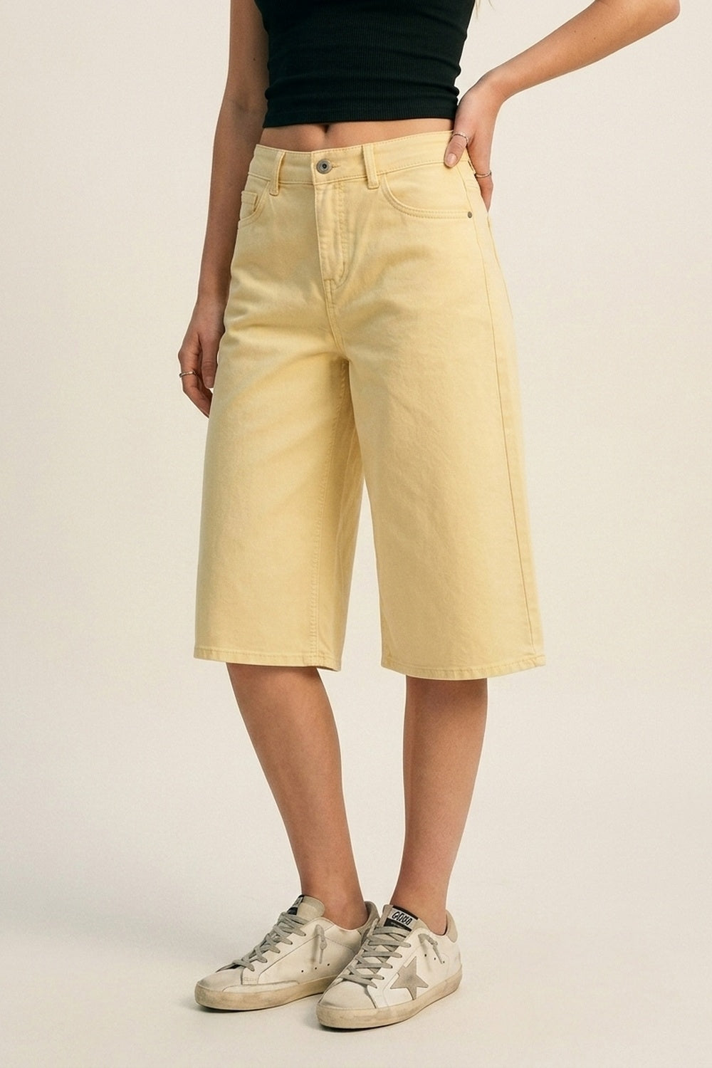 Bermudas vaqueras tipo culotte en color amarillo pastel de tiro alto, combinadas con top cropped negro y zapatillas deportivas envejecidas.