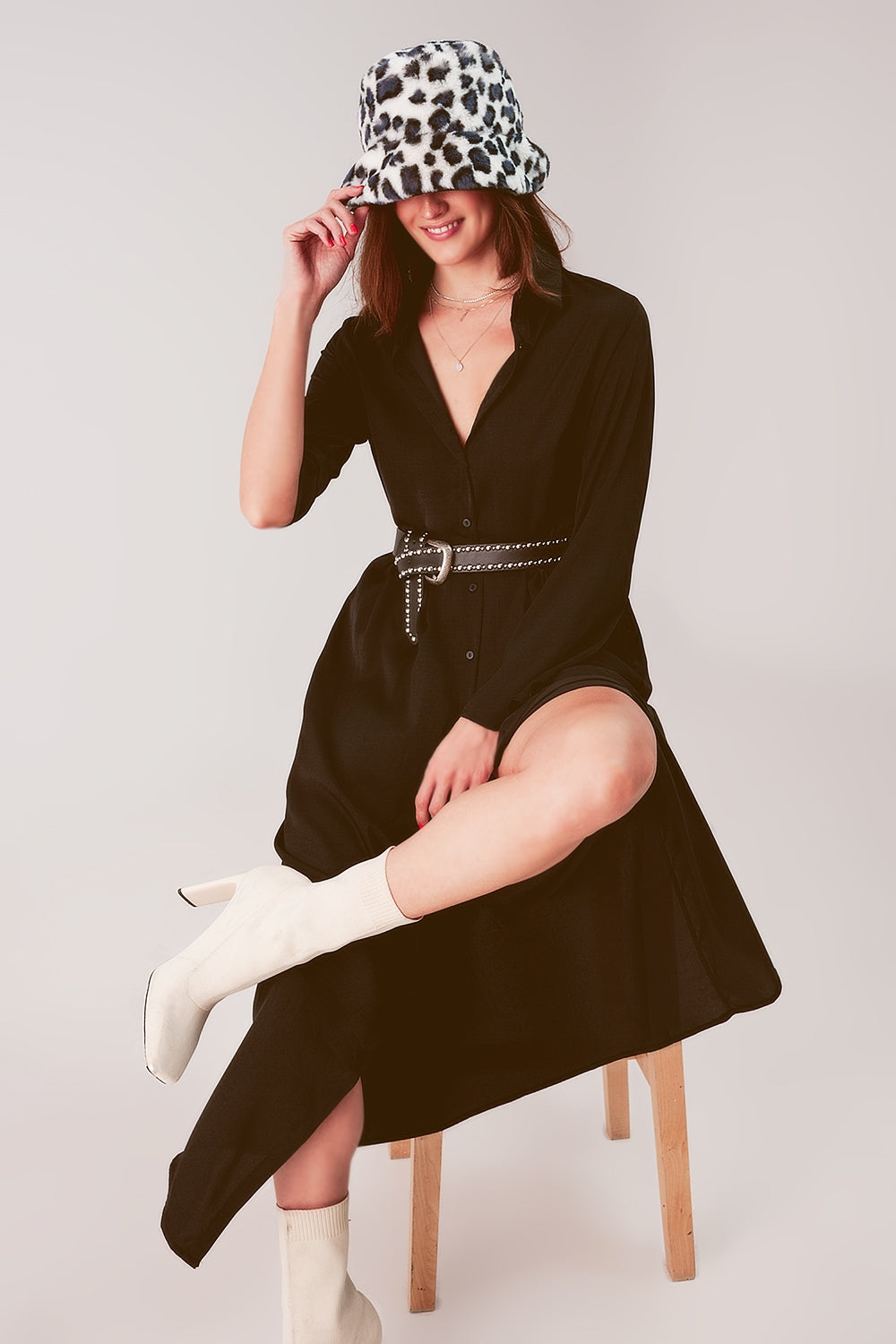 Modelo sentada con un vestido camisero largo de satén en color negro, con cinturón y botas.