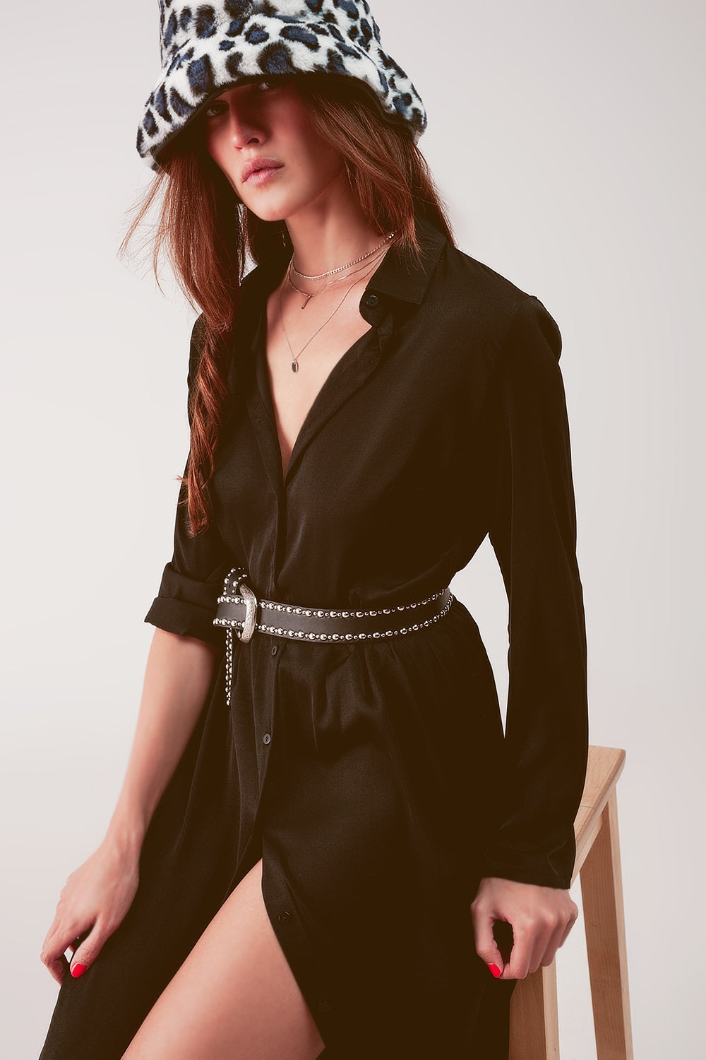 Modelo sentada con un vestido camisero largo de satén en color negro, con cinturón y botas.