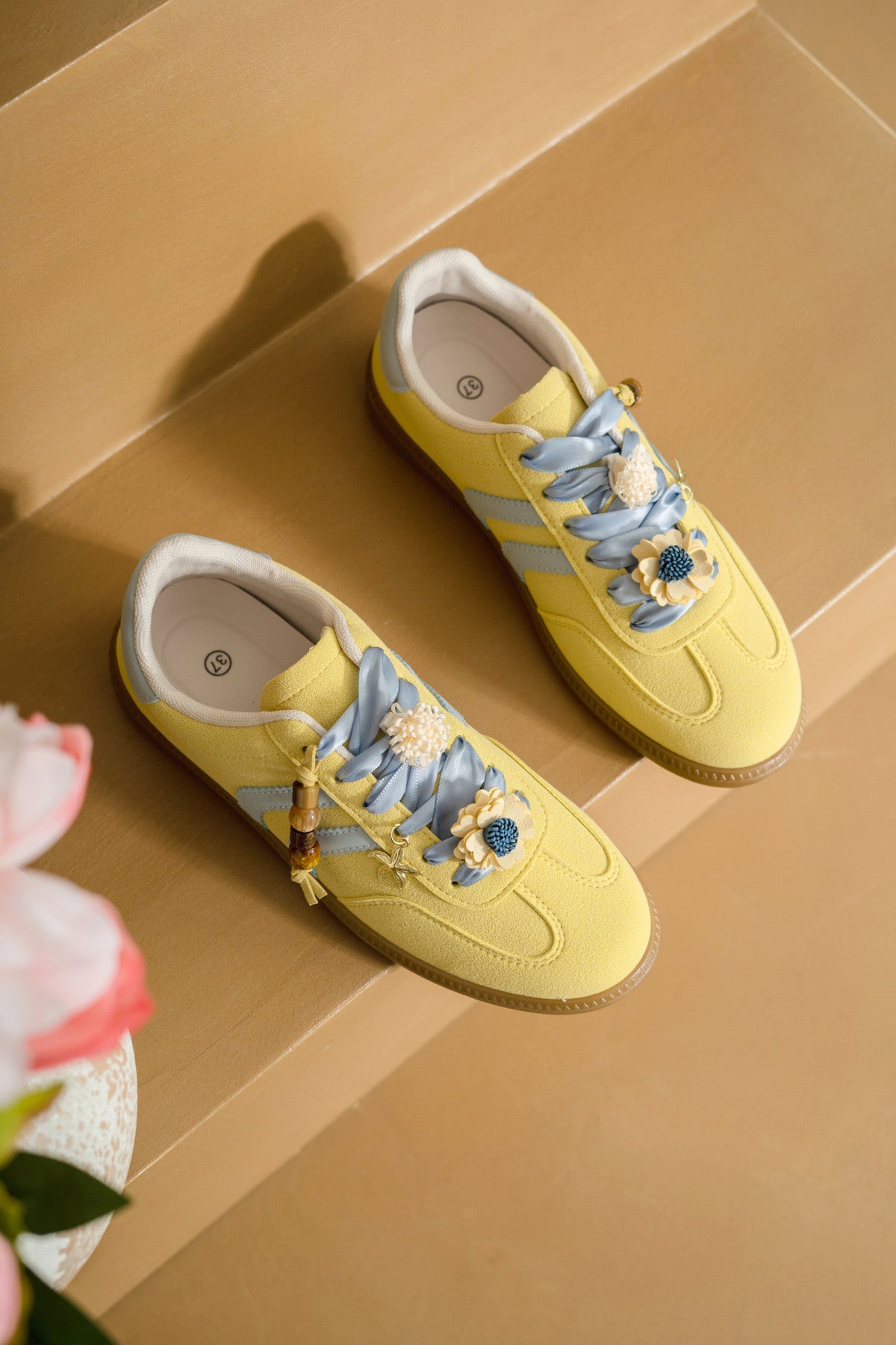 Zapatilla deportiva de mujer efecto antelina amarillo pastel con cordones celestes de satén, detalles de flores 3D y suela de goma.