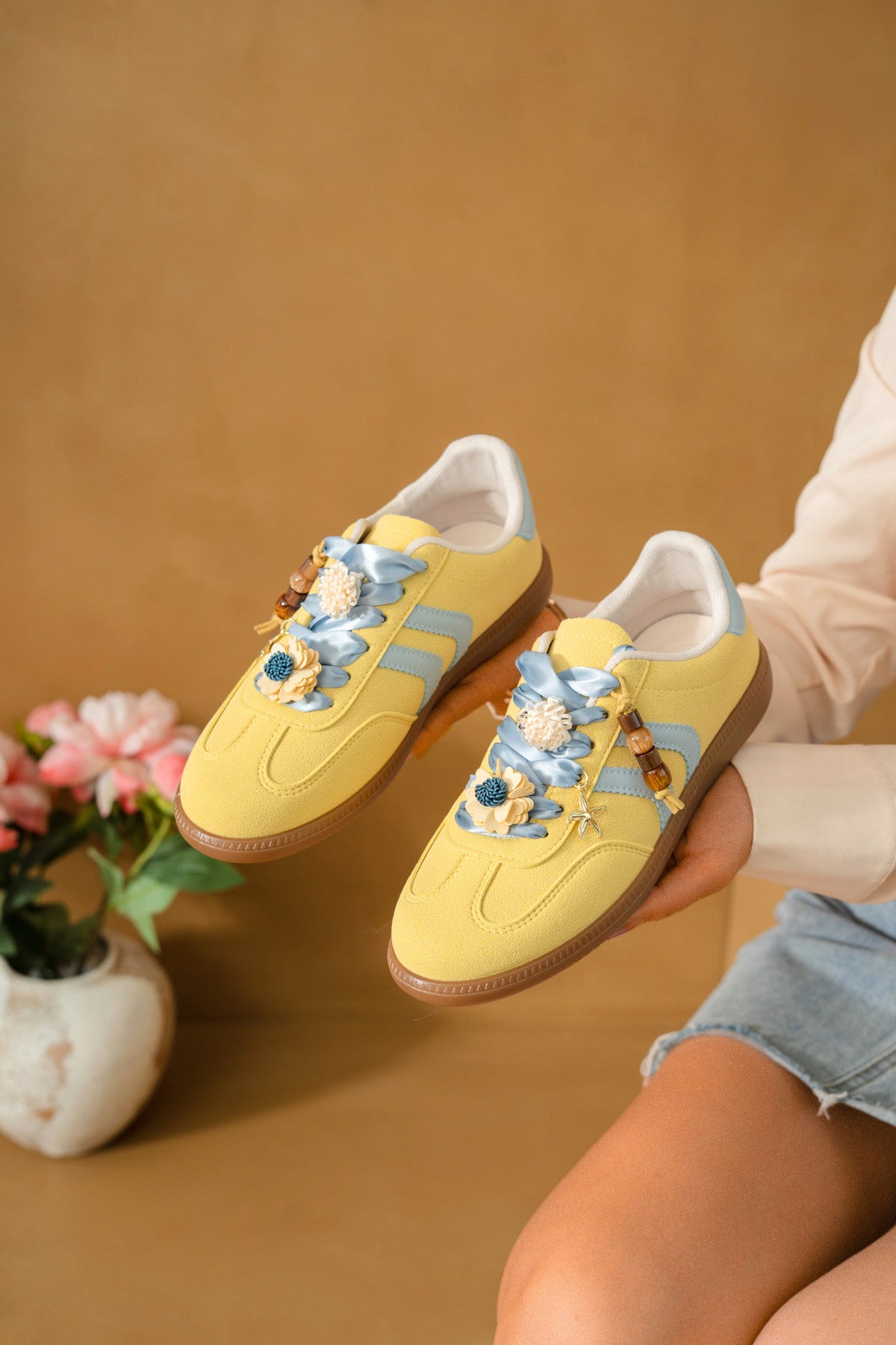 Zapatilla deportiva de mujer efecto antelina amarillo pastel con cordones celestes de satén, detalles de flores 3D y suela de goma.