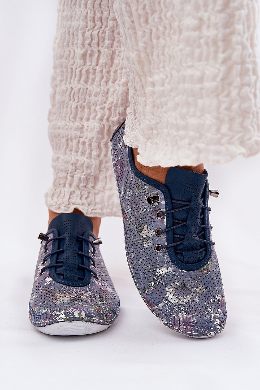 Zapatillas de piel en azul marino con estampado floral metalizado y elásticos, con pantalón blanco arrugado