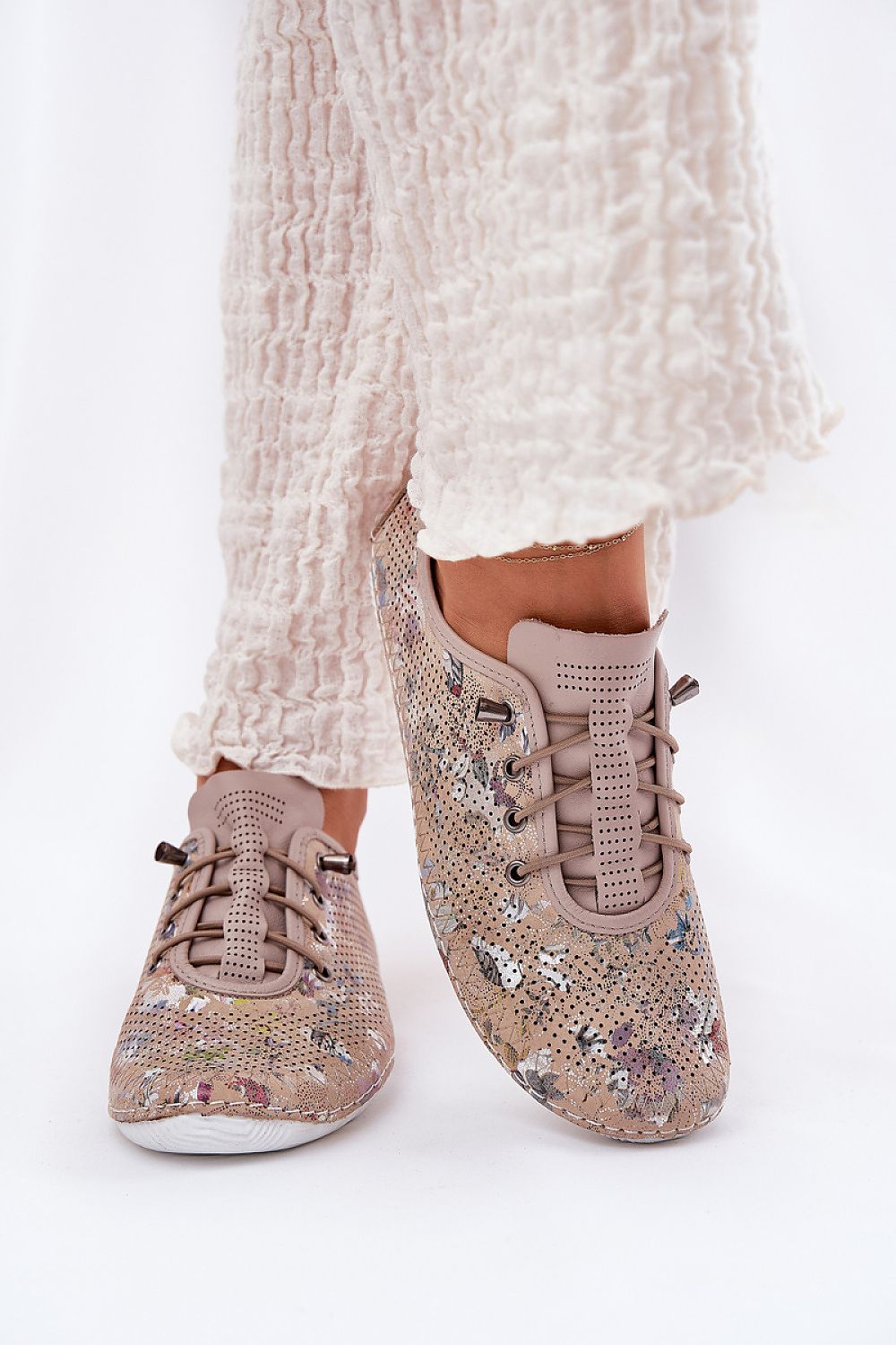 Zapatillas de piel en beige con estampado floral y elásticos simulando cordones, combinadas con pantalón blanco fruncido.
