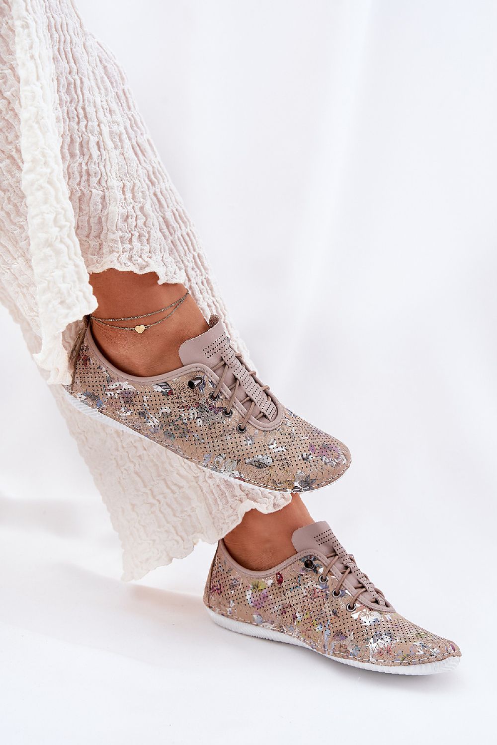 Zapatillas de piel en beige con estampado floral y elásticos simulando cordones, combinadas con pantalón blanco fruncido en movimiento