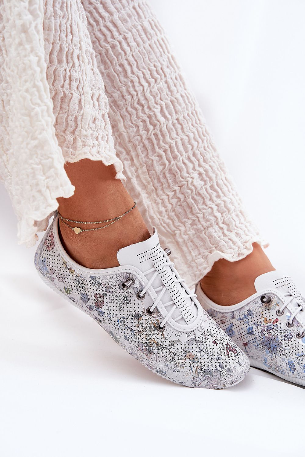 Zapatillas de piel blanca con detalles florales, vista lateral en conjunto con pantalón fluido blanco.