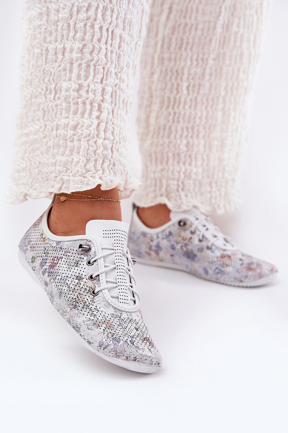 Zapatillas de piel blanca con detalles florales , vista lateral en conjunto con pantalón fluido blanco.